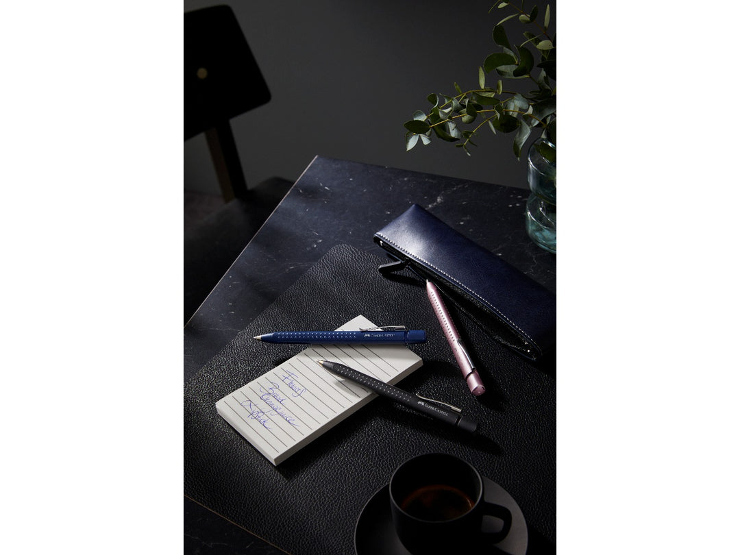 Faber-Castell Grip 2011 Ballpoint Pens in Classic Blue, Matt Black and Pale Rose displayed on a desk with a notebook and pen case, showcasing the ergonomic triangular barrel, patented soft-grip dots, metal pocket clips and smooth extra broad blue ink for everyday writing, note-taking and office use.