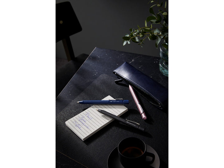 Faber-Castell Grip 2011 Ballpoint Pens in Classic Blue, Matt Black and Pale Rose displayed on a desk with a notebook and pen case, showcasing the ergonomic triangular barrel, patented soft-grip dots, metal pocket clips and smooth extra broad blue ink for everyday writing, note-taking and office use.