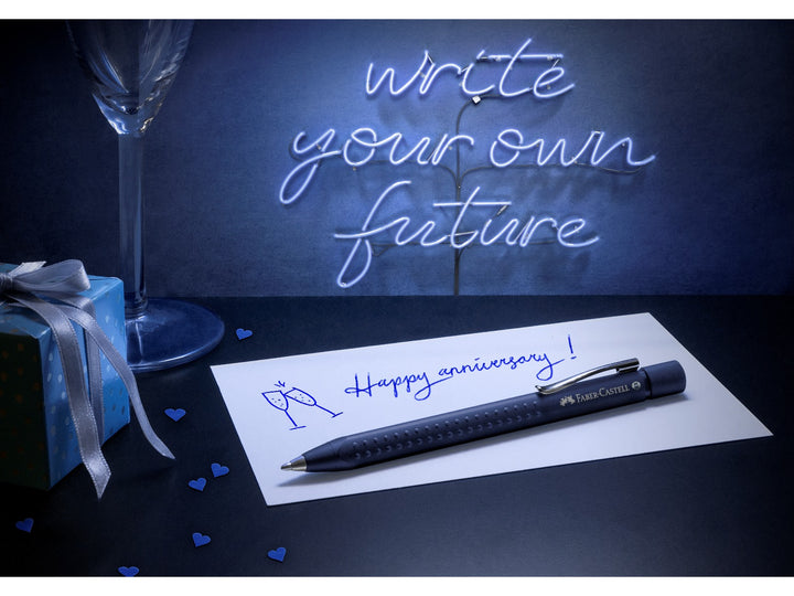 Faber-Castell Grip 2011 Ballpoint Pen in Classic Blue displayed with a handwritten anniversary card, gift box and decorative desk accessories, demonstrating the ergonomic triangular barrel, patented soft-grip dots, metal pocket clip and smooth extra broad blue ink for personal correspondence, everyday writing and gifting.