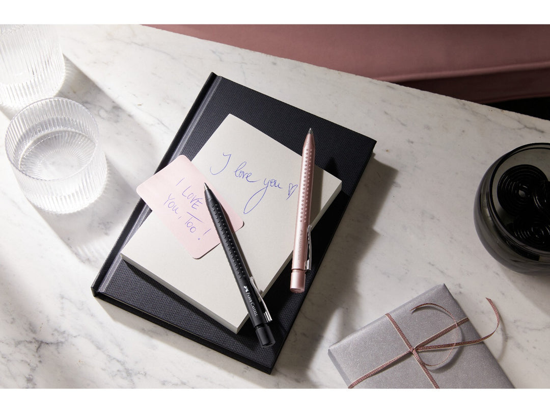 Faber-Castell Grip 2011 Ballpoint Pens in Matt Black and Pale Rose displayed on a notebook with handwritten notes, demonstrating the ergonomic triangular barrel, patented soft-grip dots, metal pocket clip and smooth extra broad blue ink for everyday writing, note-taking and office use.