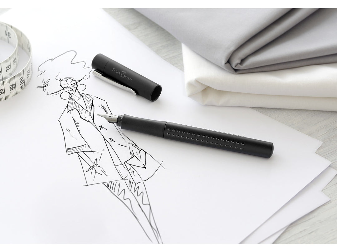 Faber-Castell Grip 2011 Fountain Pen in Black shown beside a fashion sketch and measuring tape, featuring an ergonomic triangular barrel with patented Grip dots, durable stainless steel broad nib and metal pocket clip for handwriting, fashion illustration, sketching, journaling and professional writing.
