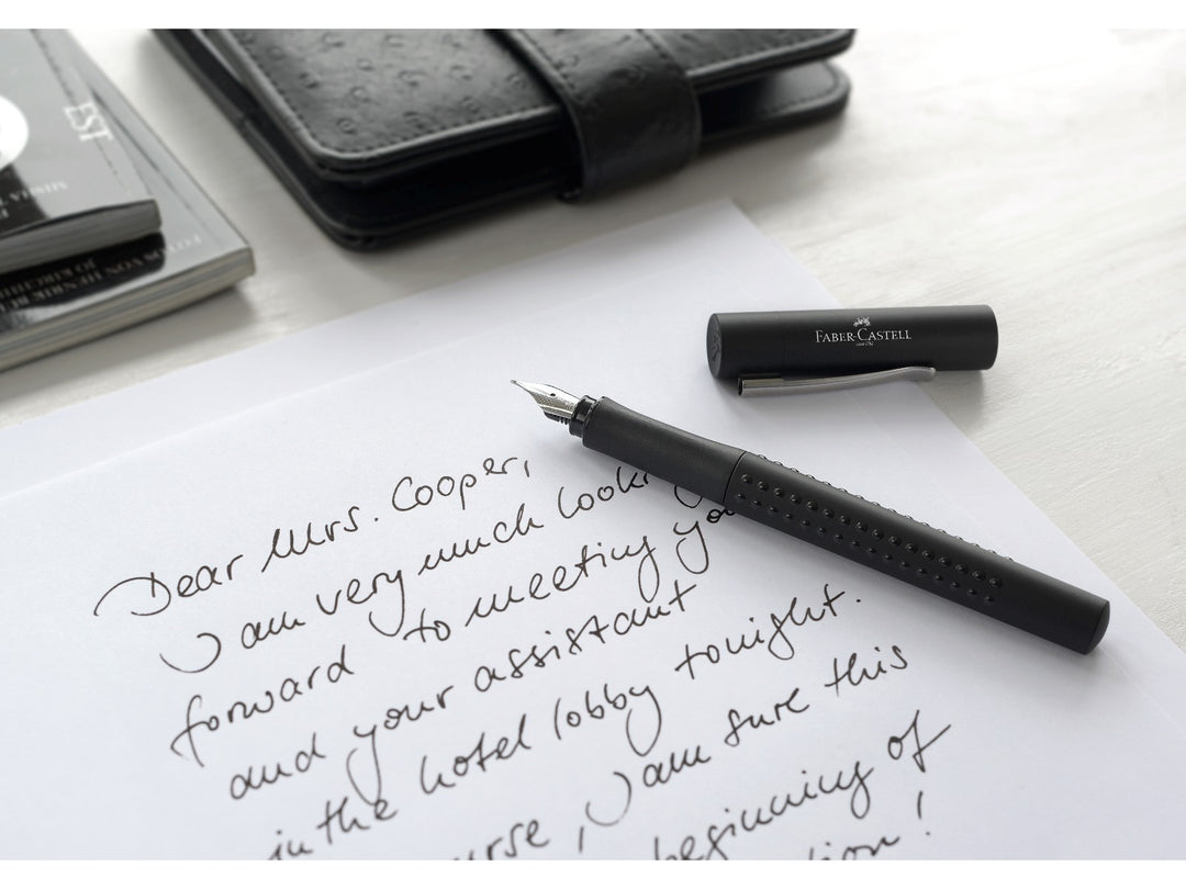Faber-Castell Grip 2011 Fountain Pen in Black shown writing a handwritten letter on white paper, featuring an ergonomic triangular barrel with patented Grip dots, durable stainless steel broad nib and metal pocket clip for comfortable everyday handwriting, journaling, note-taking and professional writing.