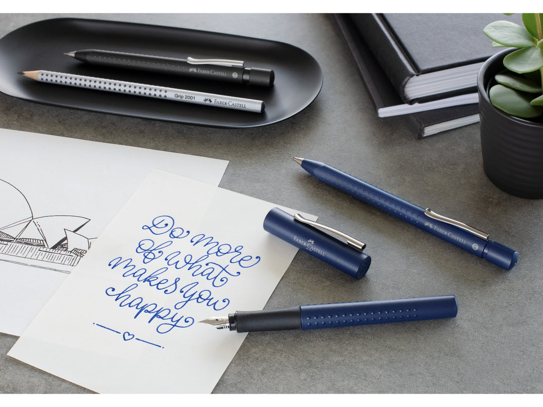 Lifestyle image featuring the Faber-Castell Grip 2011 writing collection in Classic Blue, including a fountain pen, ballpoint pen and Grip 2001 graphite pencil displayed beside handwritten notes, a sketch, notebooks and a potted plant, highlighting the ergonomic Grip dot design, comfortable writing performance and coordinated everyday stationery range.