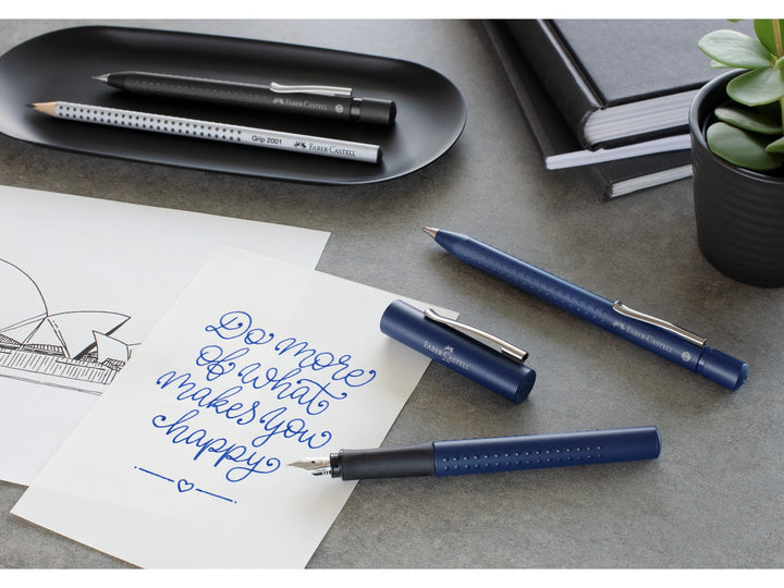 Lifestyle image featuring the Faber-Castell Grip 2011 writing collection in Classic Blue, including a fountain pen, ballpoint pen and Grip 2001 graphite pencil displayed beside handwritten notes, a sketch, notebooks and a potted plant, highlighting the ergonomic Grip dot design, comfortable writing performance and coordinated everyday stationery range.