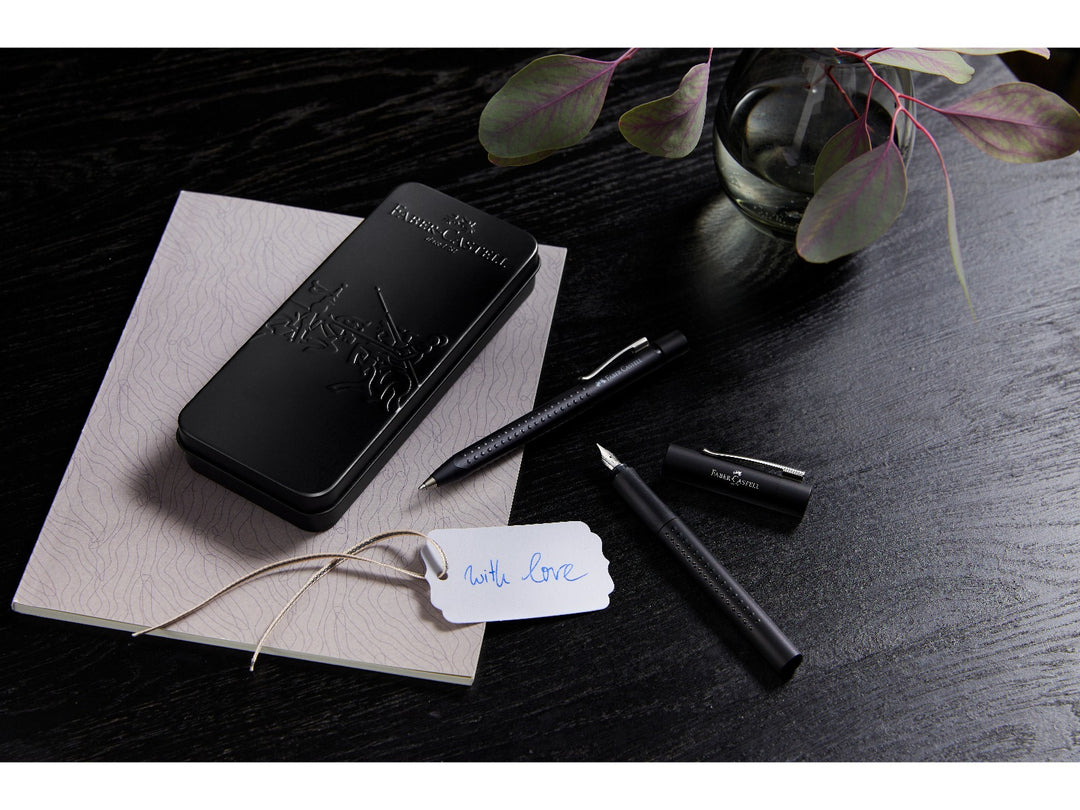 Lifestyle image featuring the Faber-Castell Grip 2011 Fountain Pen Gift Set Black 2 Pieces displayed with its premium presentation tin, matching fountain pen and ballpoint pen, shown alongside a gift tag and notebook, highlighting the ergonomic Grip dot design, stainless steel fountain pen nib and elegant gift-ready presentation for everyday writing, journaling and gifting.