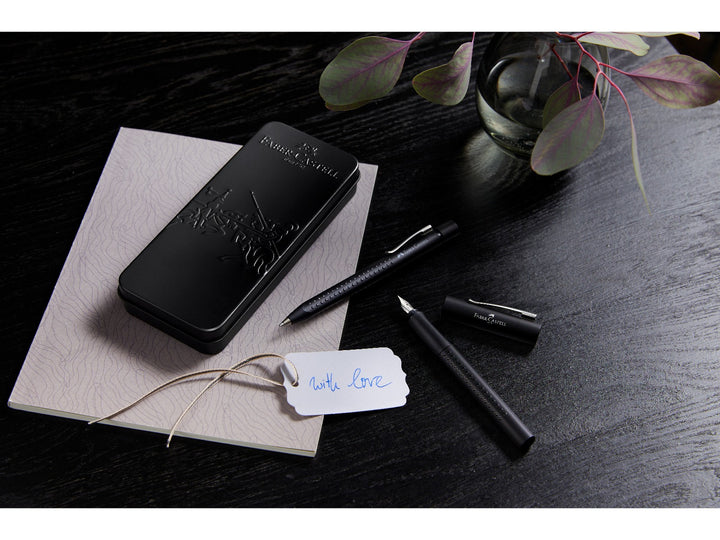 Lifestyle image featuring the Faber-Castell Grip 2011 Fountain Pen Gift Set Black 2 Pieces displayed with its premium presentation tin, matching fountain pen and ballpoint pen, shown alongside a gift tag and notebook, highlighting the ergonomic Grip dot design, stainless steel fountain pen nib and elegant gift-ready presentation for everyday writing, journaling and gifting.
