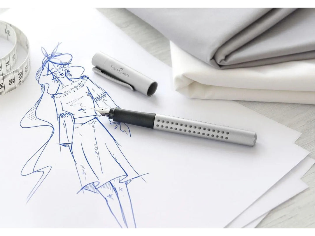 Faber-Castell Grip 2011 Fountain Pen in Silver shown beside a fashion illustration and measuring tape, featuring an ergonomic triangular barrel with patented Grip dots, durable stainless steel nib and metal pocket clip for handwriting, fashion sketching, journaling, note-taking and creative design work.