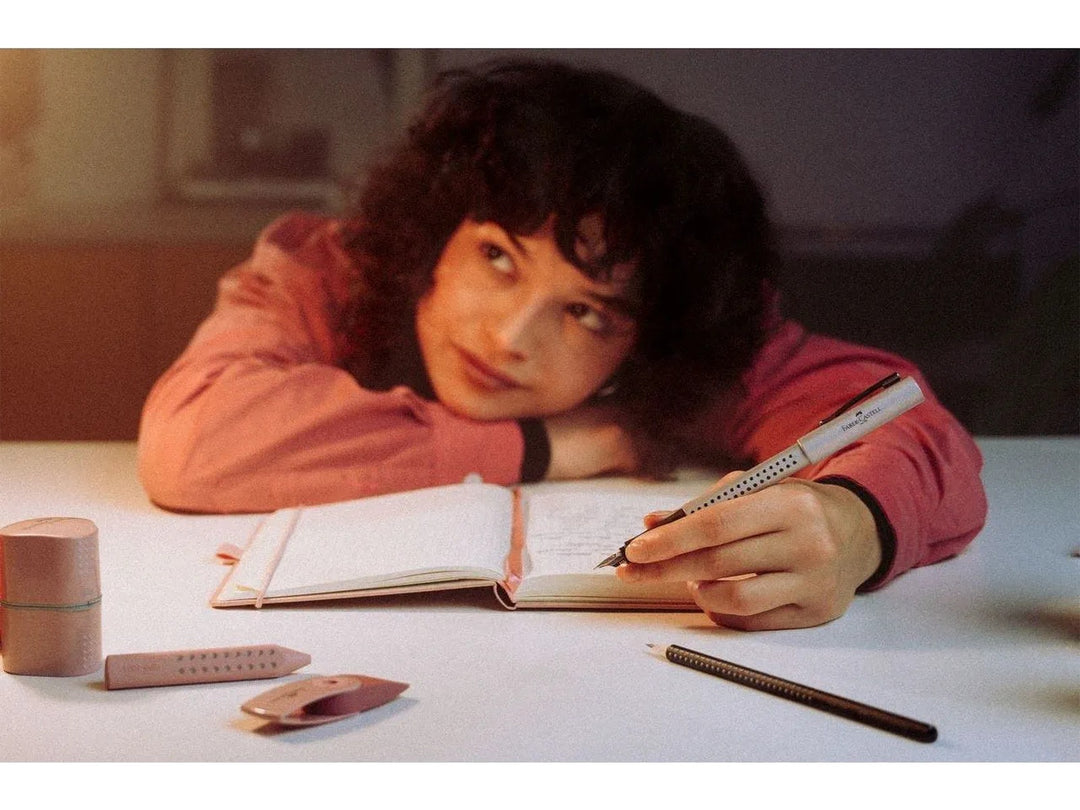 Lifestyle image featuring the Faber-Castell Grip Edition Glam Fountain Pen in Pearl writing in an open notebook, displayed alongside matching Glam writing instruments, books and a decorative neon sign, highlighting the shimmering pearlescent finish, ergonomic Grip dot design, stainless steel nib and smooth writing performance for journaling, studying and everyday handwriting.