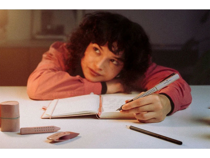Lifestyle image featuring the Faber-Castell Grip Edition Glam Writing Set Pearl in use, showing the pearl-effect fountain pen writing in an open notebook alongside the matching ballpoint pen, pencil and accessories, highlighting the ergonomic Grip dot design, coordinated writing collection and elegant pearlescent finish for journaling, study, planning and everyday writing.