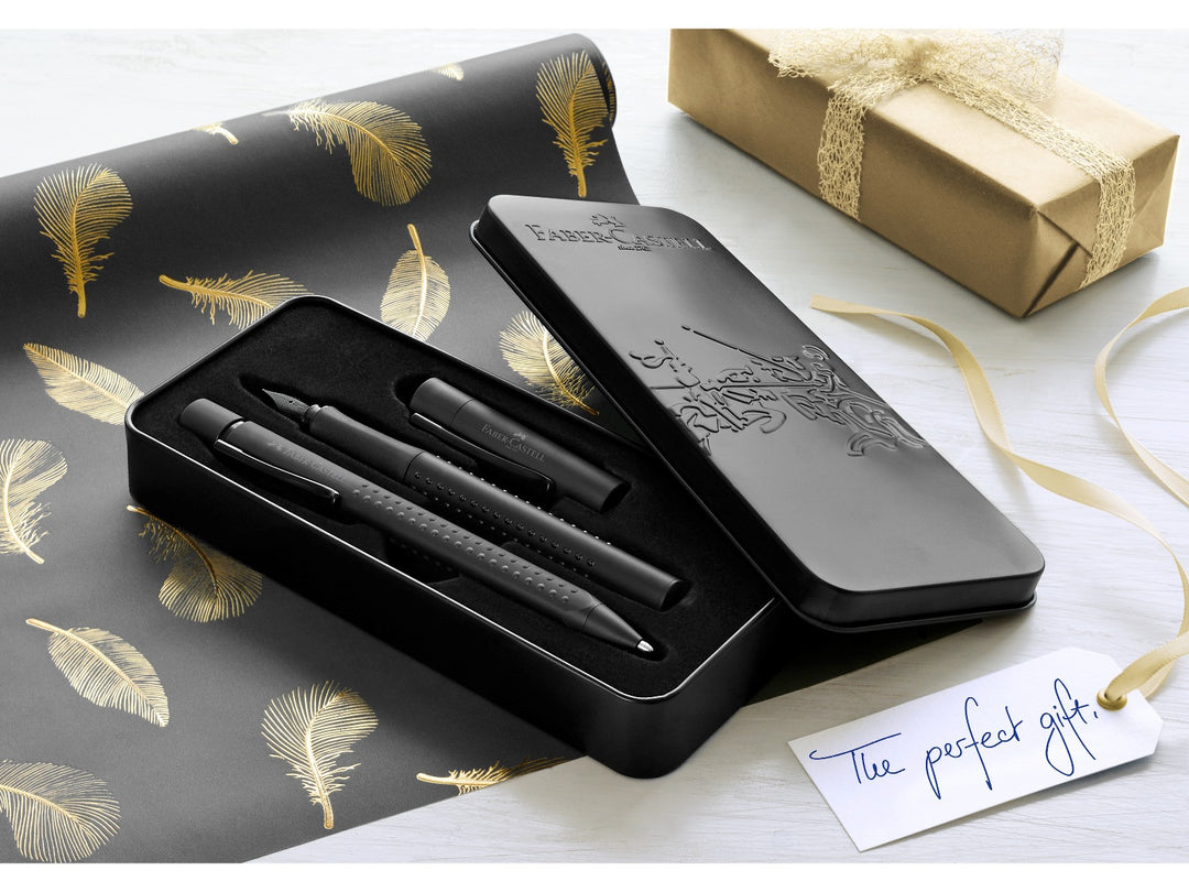 Lifestyle image featuring the Faber-Castell Grip Edition Writing Gift Set Black presented in an open premium metal gift tin, showing matching black fountain pen and ballpoint pen with ergonomic Grip dot barrels, displayed alongside decorative gift wrap, ribbon and a gift tag, highlighting the elegant presentation and suitability for gifting, journaling, correspondence and everyday writing.