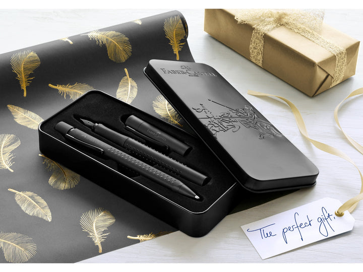 Lifestyle image featuring the Faber-Castell Grip Edition Writing Gift Set Black presented in an open premium metal gift tin, showing matching black fountain pen and ballpoint pen with ergonomic Grip dot barrels, displayed alongside decorative gift wrap, ribbon and a gift tag, highlighting the elegant presentation and suitability for gifting, journaling, correspondence and everyday writing.