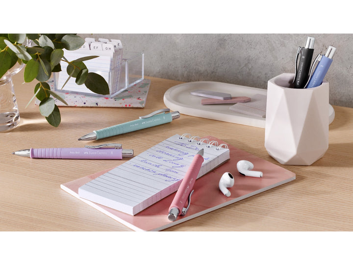 Lifestyle scene featuring Faber-Castell Poly Ball ballpoint pens in Rose, Mint Green, Lilac, Black and Light Blue displayed on a desk with a notepad, pen holder, index cards and wireless earbuds, highlighting the ergonomic soft-touch grip zones, refillable design and smooth XB writing performance for everyday writing and note-taking.