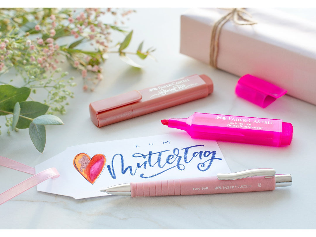Lifestyle scene featuring a Faber-Castell Poly Ball Ballpoint Pen XB in Rose alongside pastel stationery, a fluorescent pink highlighter and a handwritten gift tag, highlighting the ergonomic soft-touch grip zone, refillable design and smooth extra broad writing performance for everyday writing, journaling and personal correspondence.
