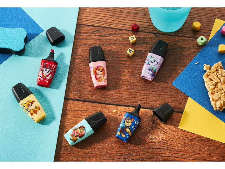 Six STABILO BOSS MINI PAW Patrol highlighters featuring colourful character designs arranged across a wooden desk with paper, letter blocks and craft materials