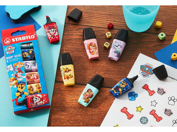 Six STABILO BOSS MINI PAW Patrol highlighters arranged on a wooden craft table alongside the retail wallet and PAW Patrol activity sheet, showing the colourful character designs in use