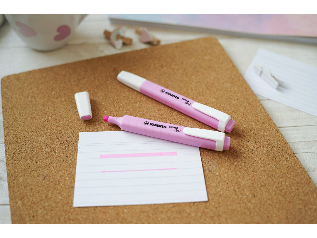 Pink STABILO swing cool highlighters shown capped and uncapped on a cork desk mat, demonstrating the chisel tip with broad and fine pink highlighting lines on note paper