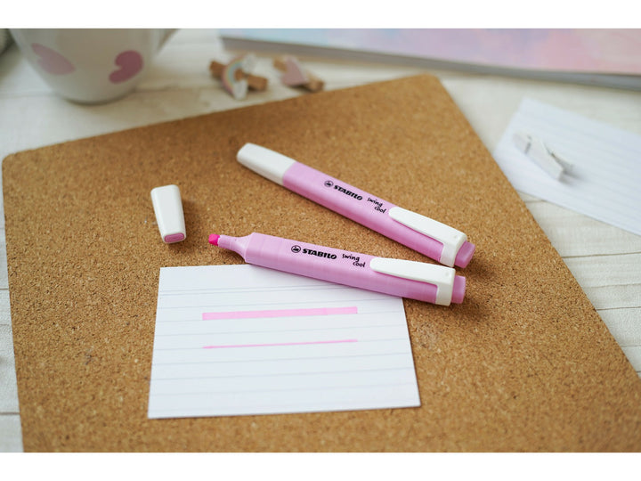 Pink STABILO swing cool highlighters shown capped and uncapped on a cork desk mat, demonstrating the chisel tip with broad and fine pink highlighting lines on note paper