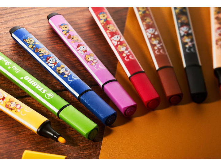 Eight STABILO Trio Scribbi PAW Patrol felt-tip pens in assorted colours arranged on a wooden desk, featuring colourful PAW Patrol character designs and triangular barrels