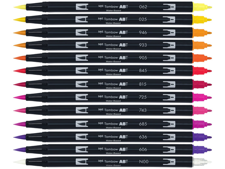 Tombow ABT Dual Brush Pen Sunset Colours Set of 12 displayed individually, featuring Pale Yellow (062), Light Orange (025), Gold Ochre (946), Orange (933), Red (905), Carmine (845), Cherry (815), Rhodamine Red (725), Hot Pink (743), Deep Magenta (685), Imperial Purple (636), Violet (606) and a colourless Blender (N00), each with dual brush and fine tips visible for hand lettering, colouring, illustration and watercolour-style effects.