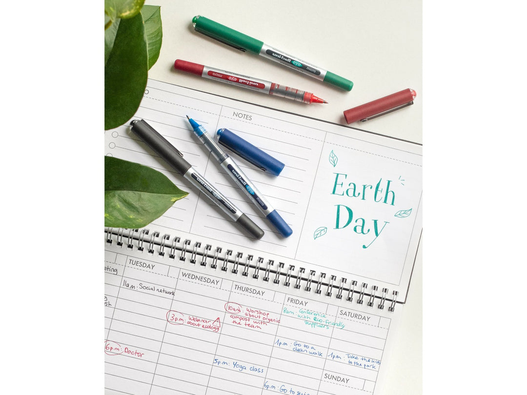 uni-ball Eye Micro UB-150 rollerball pens in Black, Blue, Green and Red displayed on a desk planner, showcasing visible ink reservoirs, precision micro tips and smooth liquid Super Ink writing performance