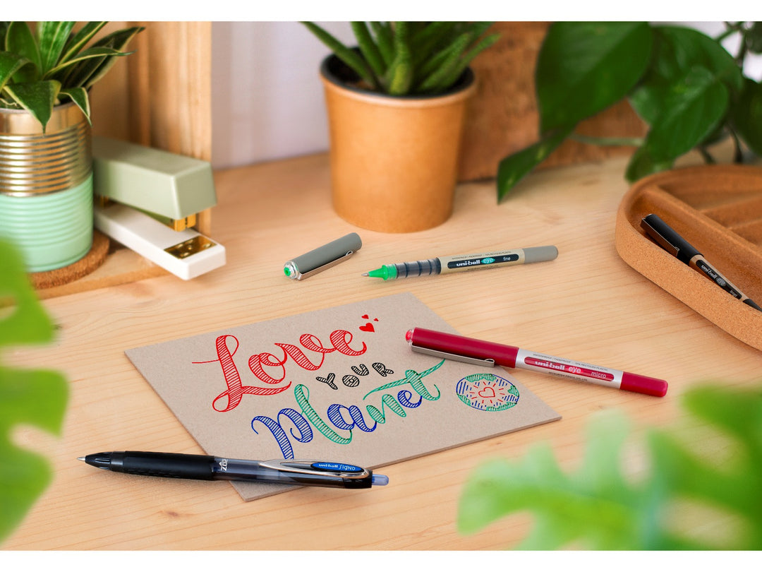 uni-ball Eye rollerball pens in Black, Green and Wine displayed on a desk with a handwritten card, showcasing smooth liquid Super Ink writing, visible ink reservoirs and everyday writing performance