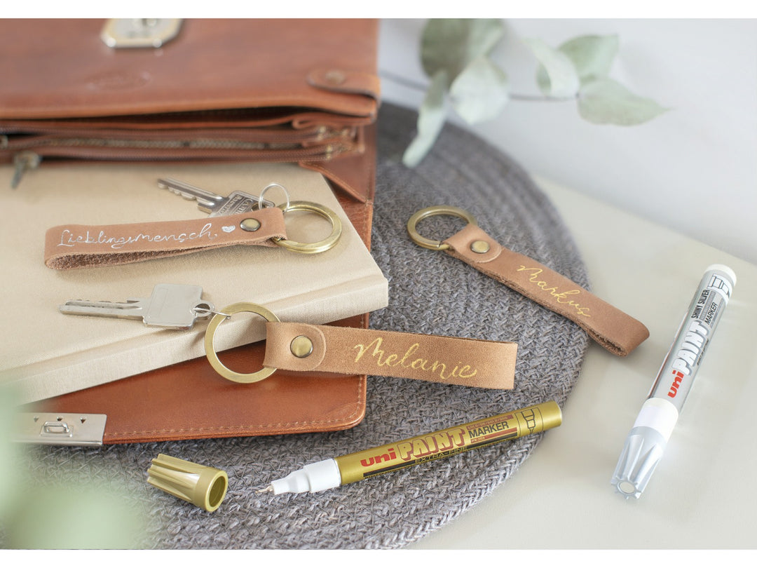 Gold and white uni-ball PX-203 extra fine paint markers displayed with personalised leather keyrings, showing metallic gold and white writing on brown leather