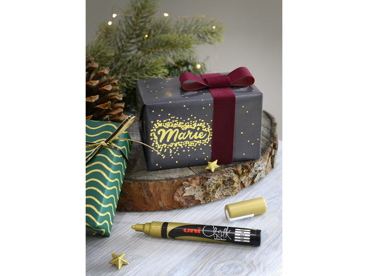 Gold uni ChalkGlass marker displayed beside a personalised Christmas gift, showing metallic gold lettering and decorative details on dark gift wrap