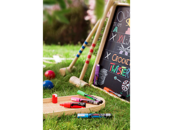 Lifestyle image showing uni ChalkGlass PWE-8K liquid chalk markers being used outdoors on a chalkboard scoreboard during a garden game, featuring colourful wipe-clean lettering and illustrations alongside fluorescent chalk markers, demonstrating use for event signs, promotional displays, menu boards, celebrations and creative writing on non-porous surfaces such as chalkboards and glass.