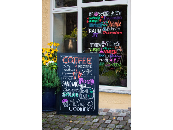 Lifestyle image showing uni ChalkGlass PWE-8K liquid chalk markers used to create colourful café menu boards and window displays, featuring bold wipe-clean lettering and illustrations on a freestanding chalkboard sign and shop window, demonstrating suitability for restaurant menus, retail signage, promotional displays and creative hand lettering on non-porous surfaces such as glass and chalkboards.