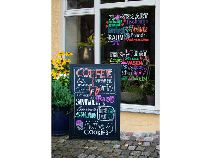 Lifestyle image showing uni ChalkGlass PWE-8K liquid chalk markers used to create colourful café menu boards and window displays, featuring bold wipe-clean lettering and illustrations on a freestanding chalkboard sign and shop window, demonstrating suitability for restaurant menus, retail signage, promotional displays and creative hand lettering on non-porous surfaces such as glass and chalkboards.