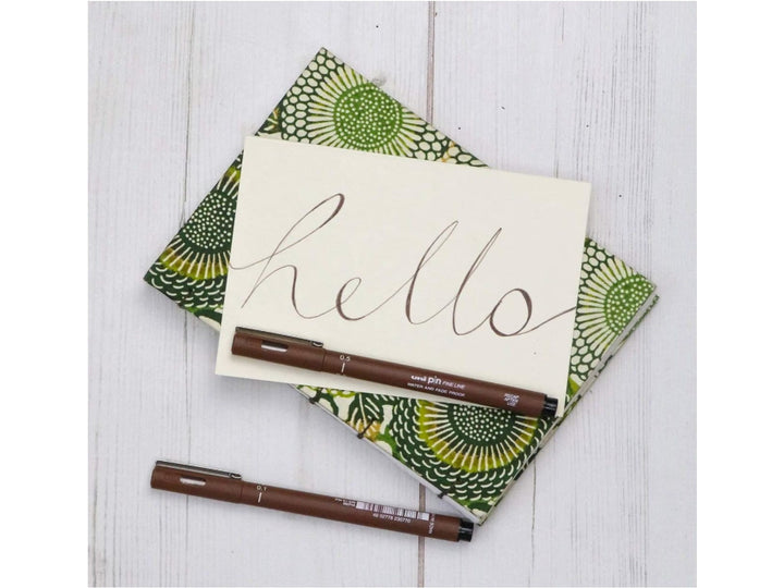 uni PIN Sepia brush pen and fineliner displayed beside a handwritten greeting card with sepia calligraphy, showcasing flexible brush lettering and archival-quality sepia pigment ink