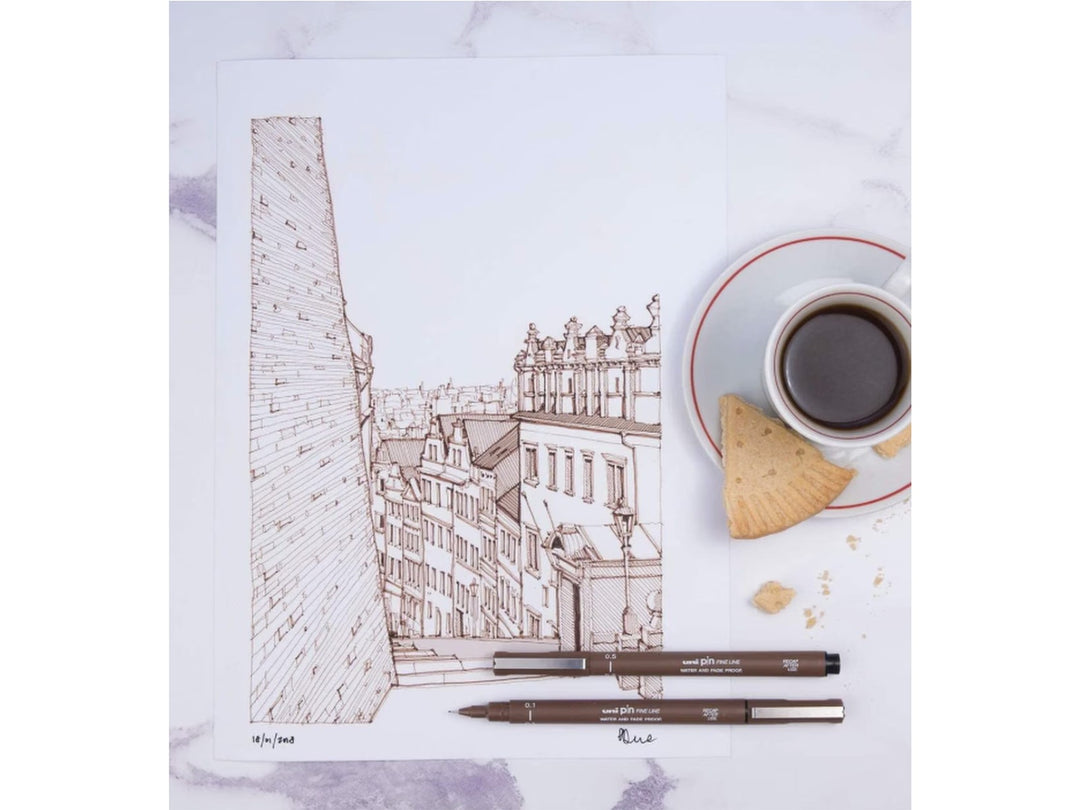 Sepia illustration created with uni PIN fineliners showing a detailed cityscape sketch alongside uni PIN Sepia 0.1mm, 0.5mm and brush pens on a desk beside a cup of coffee