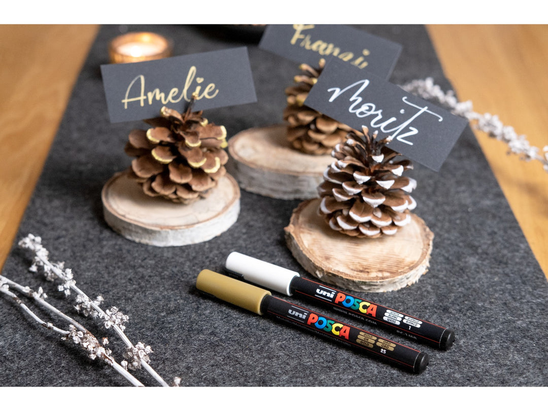 Lifestyle image showing uni POSCA PC-1MR ultra-fine paint markers in White and Gold used to create handwritten place cards on black card, displayed with decorative pine cones, wooden table decorations and dried flowers, demonstrating the 0.7mm fibre tip, opaque water-based pigment paint and precision detailing capabilities for card making, event stationery, craft projects, hand lettering and creative decoration.