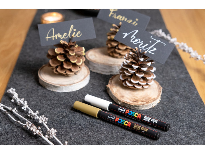 Lifestyle image showing uni POSCA PC-1MR ultra-fine paint markers in White and Gold used to create handwritten place cards on black card, displayed with decorative pine cones, wooden table decorations and dried flowers, demonstrating the 0.7mm fibre tip, opaque water-based pigment paint and precision detailing capabilities for card making, event stationery, craft projects, hand lettering and creative decoration.