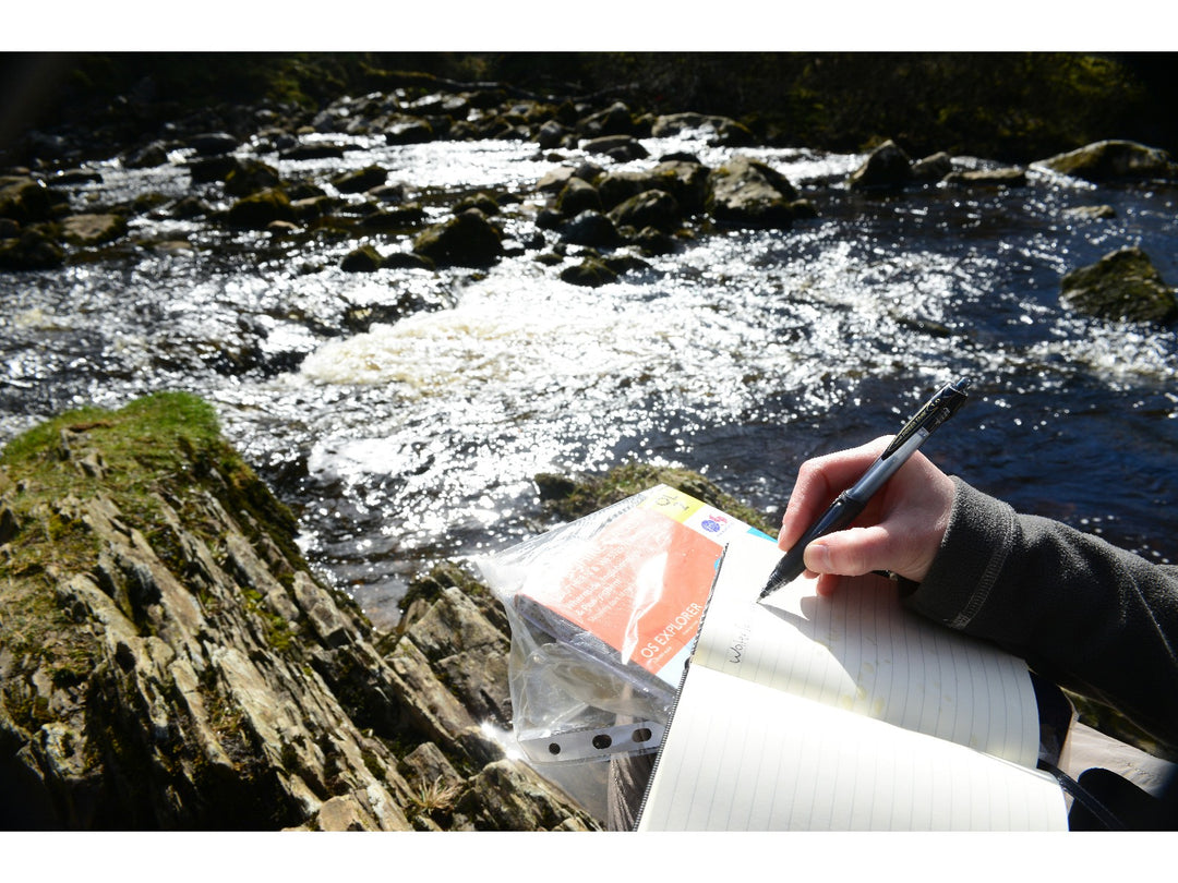 uni Power Tank SN-220 pressurised ballpoint pen being used outdoors beside a river, demonstrating reliable writing performance in challenging environments with a notebook and waterproof map case