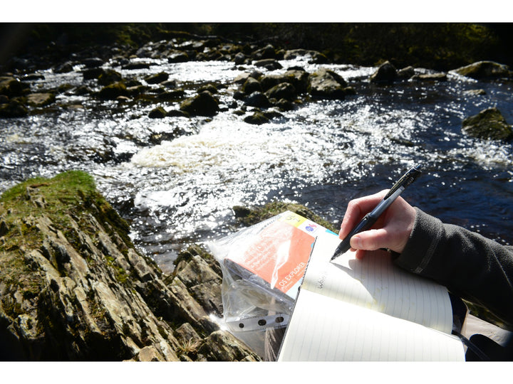 uni Power Tank SN-220 pressurised ballpoint pen being used outdoors beside a river, demonstrating reliable writing performance in challenging environments with a notebook and waterproof map case